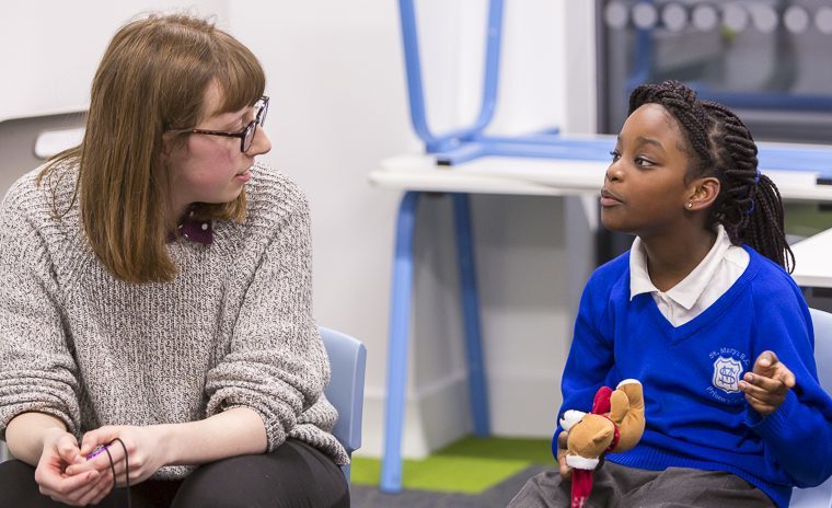A female teacher and a girl pupil sitting down inside a classroom. The pupil is speaking to the teacher and holding a little teddy bear in her hand.