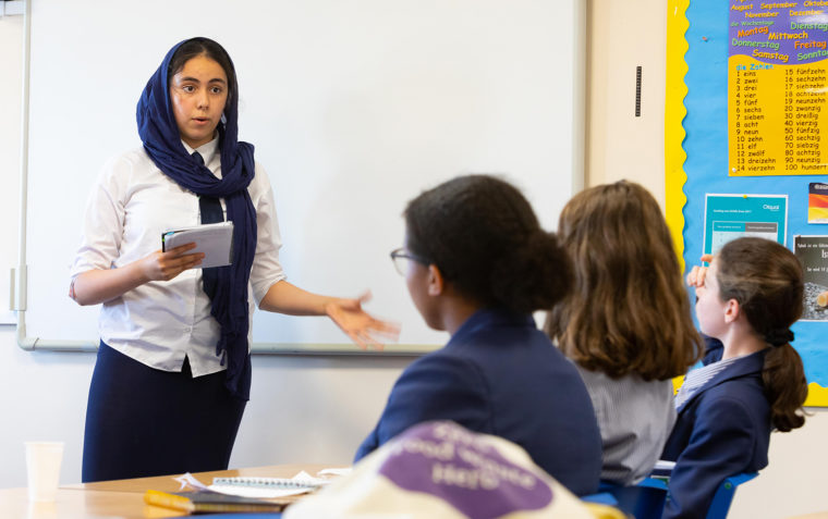 A female teacher with headscarf on standing in front of the classroom while speaking to three female students sitting and facing the blackboard.