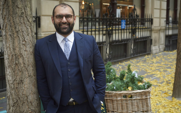 A photo of Bilal Mahmood smiling with contact lens glasses and leaning next to a tree outside a building.