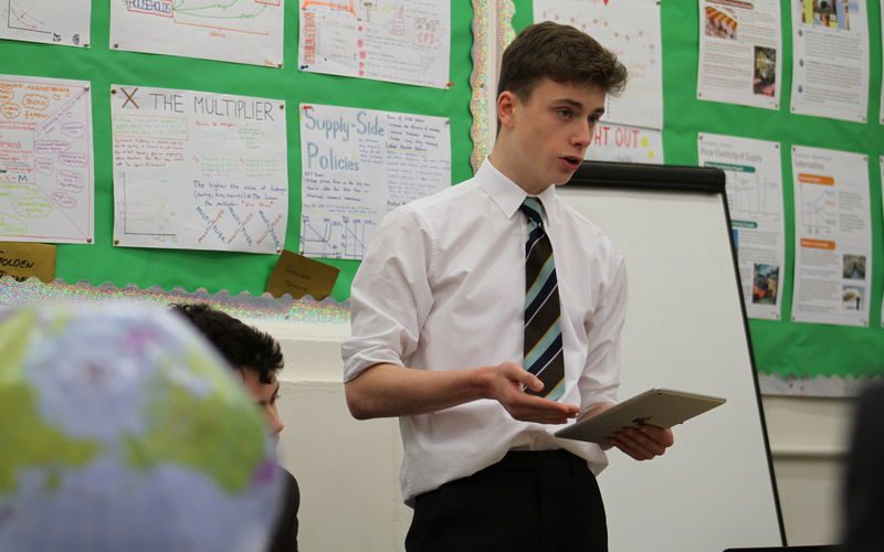 A male secondary school student standing in the classroom while reading from the notepad in his hand.