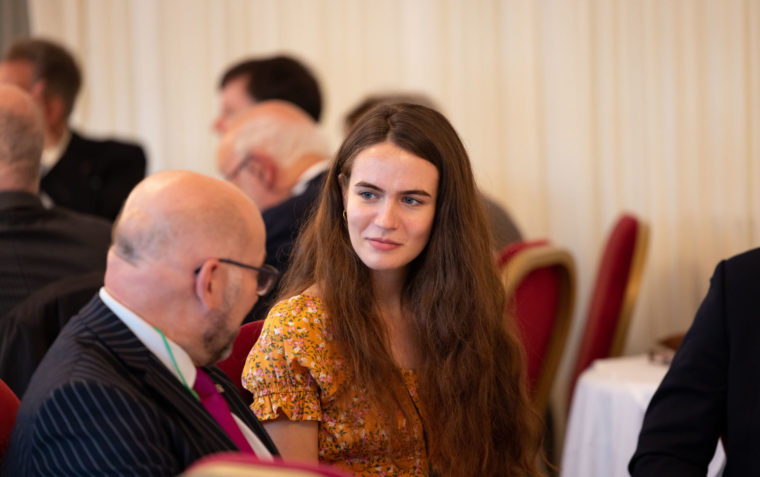 A young lady and an older man sitting inside a conference room with blurry images of other people in the background.