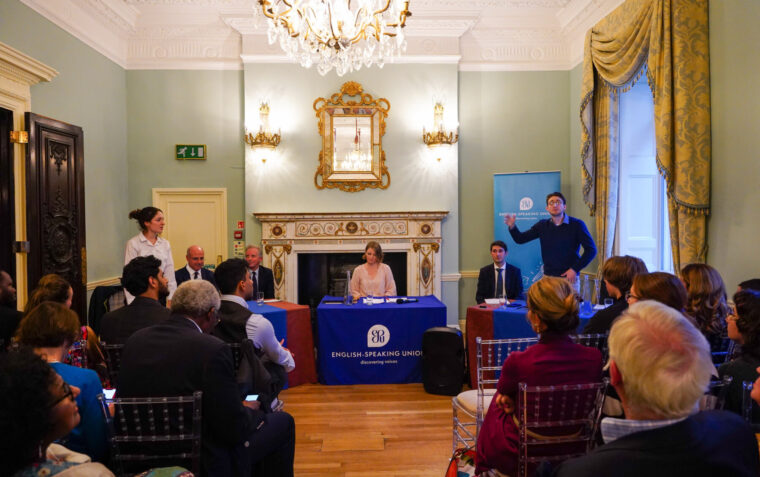 Photo of a debating event with one speaker delivering their speech while another is standing up to ask them a question