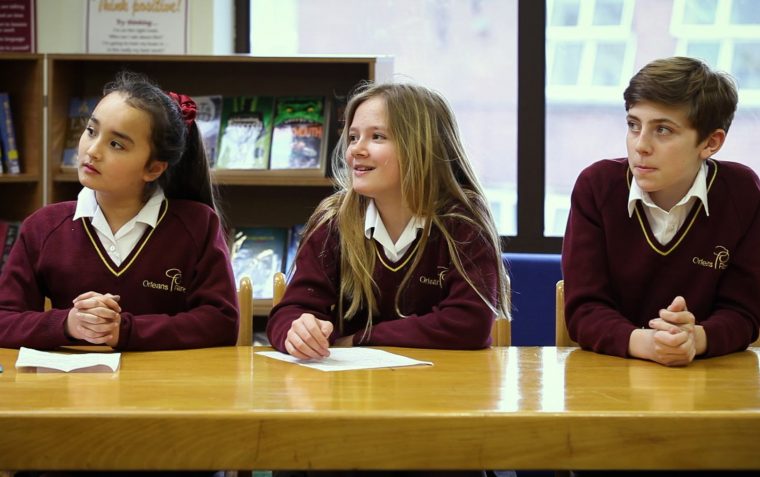Three primary school pupils (a boy and two girls) sitting down and leaning forward on the desk inside a classroom.