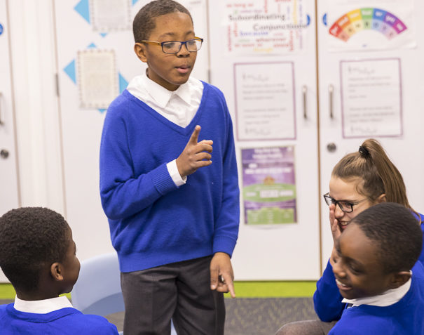 Four pupils (three boys and a girl) of St Mary’s Roman Catholic Primary School in Battersea inside a classroom. One boy is standing, and the others are sitting.