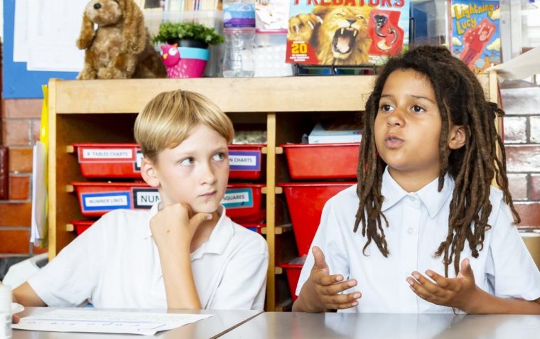 Joah and Makeda sitting down inside their classroom and Makeda is speaking.