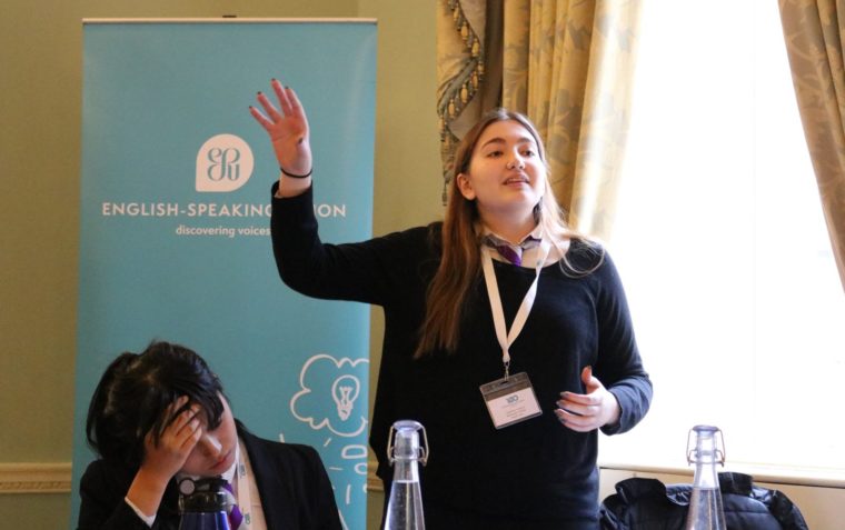 A young pupil passionately delivers her speech while her team mate is carefully listening and writing her notes