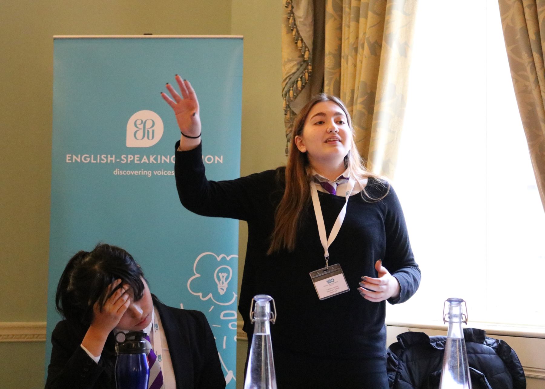 A young pupil passionately delivers her speech while her team mate is carefully listening and writing her notes