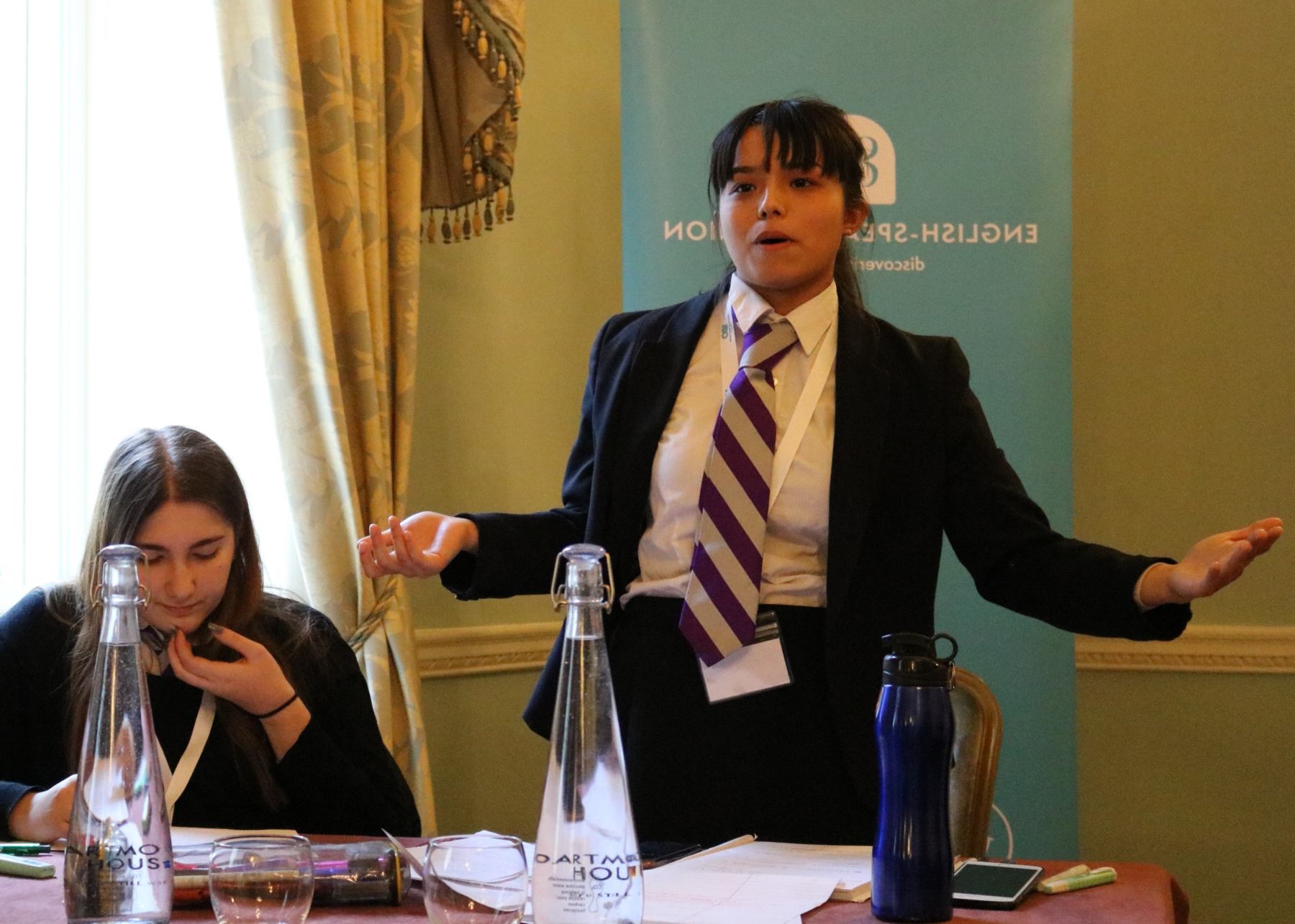 A young pupil passionately delivers her speech while her team mate is thinking and sorting her notes
