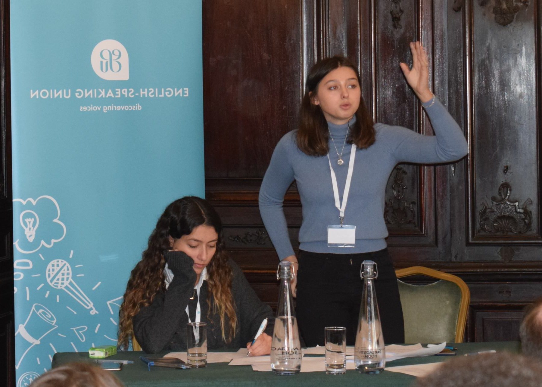 A young pupil passionately delivers her speech while her team mate is writing down her notes