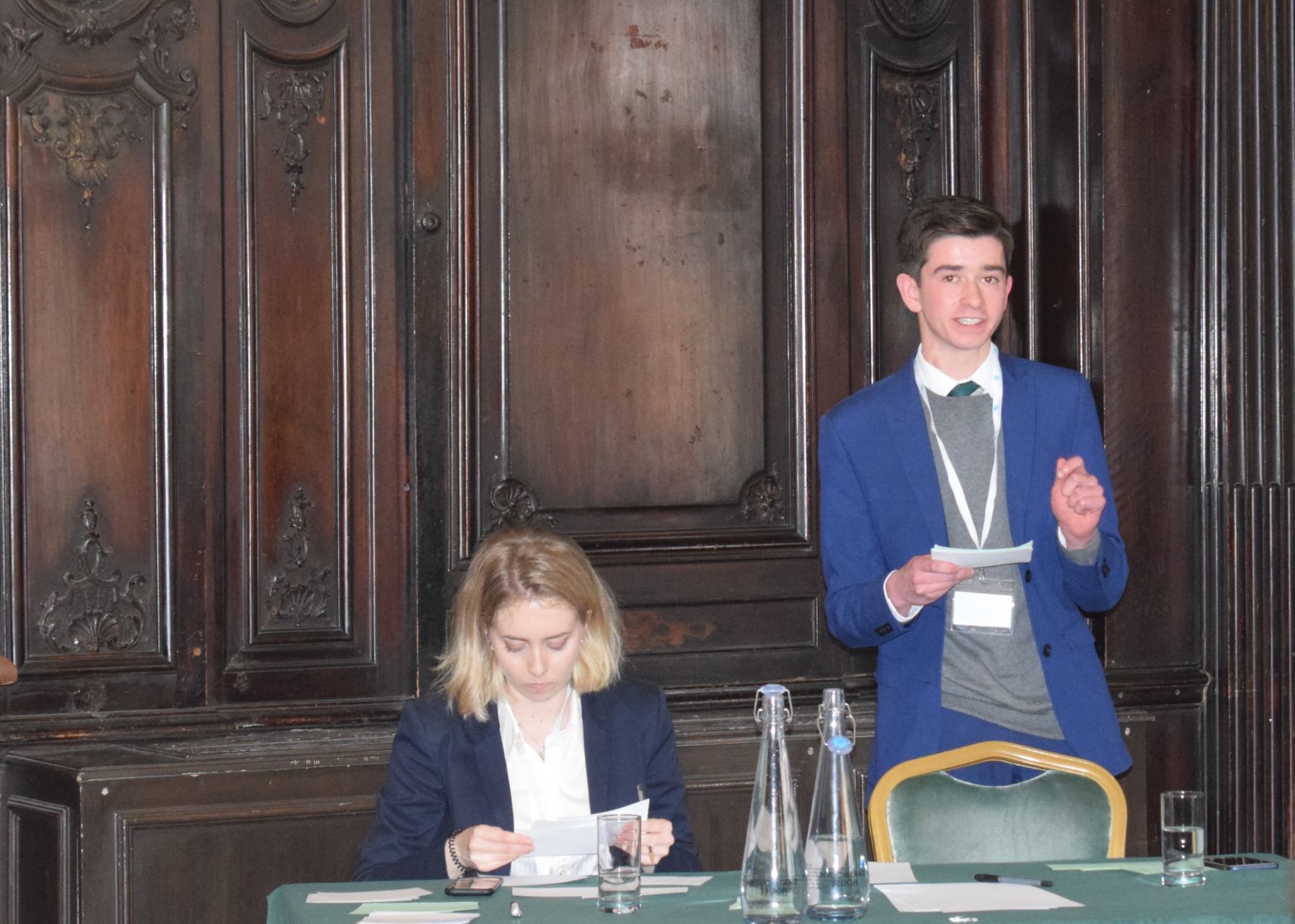 A young pupil passionately delivers his speech while his team mate is sorting out her notes