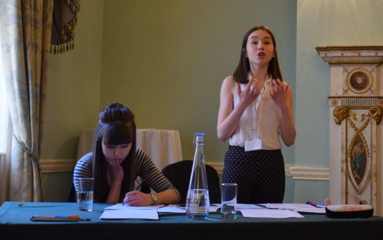 A young pupil passionately delivers her speech while her team mate is writing down her notes