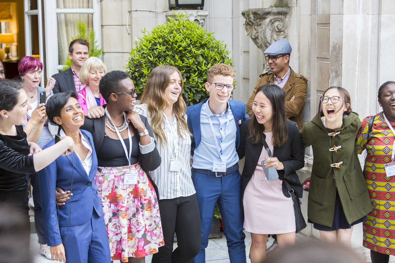 A group of secondary school students (male and female) standing outside a building smiling and laughing with hands across each other.