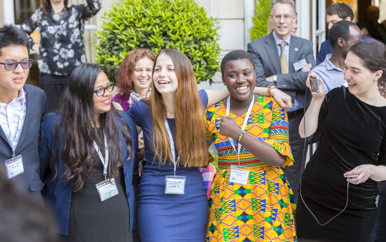 A group of international secondary school students dressed in different attires standing closed together outside a building and smiling.