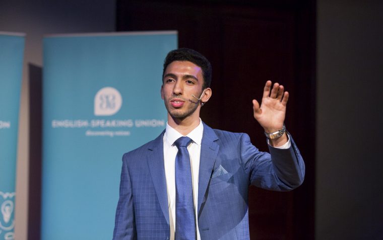 A secondary school student debater is standing and left hand stretch out while speaking at a public speaking event.