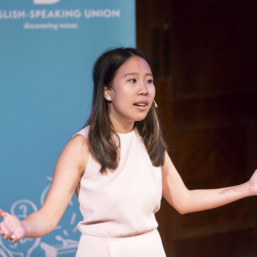 A young female secondary school student is speaking on stage with both hands spread out wide.