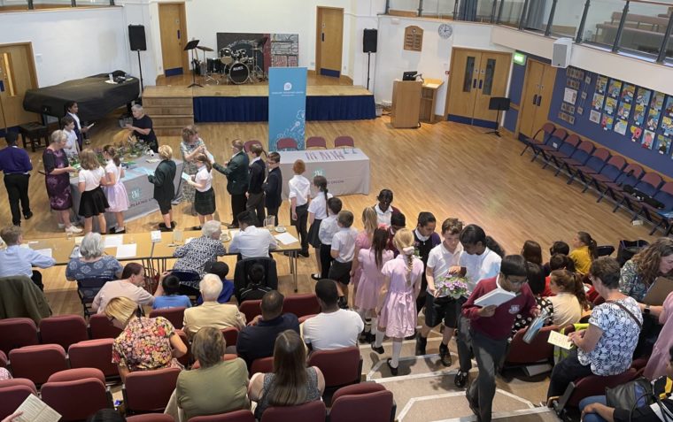 An indoor image of a theatre with school children climbing to their seats, other collecting booklets from a registration table at the centre and parents seating.