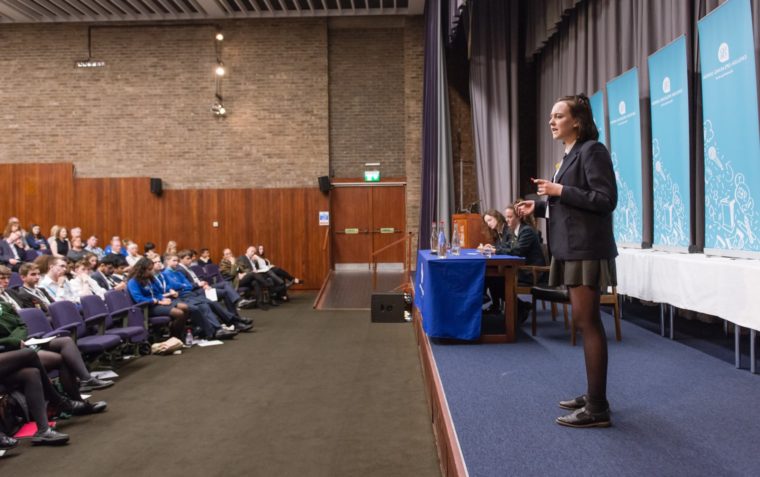 Young pupil in school uniform standing up and speaking on stage, two other pupils are on stage with her