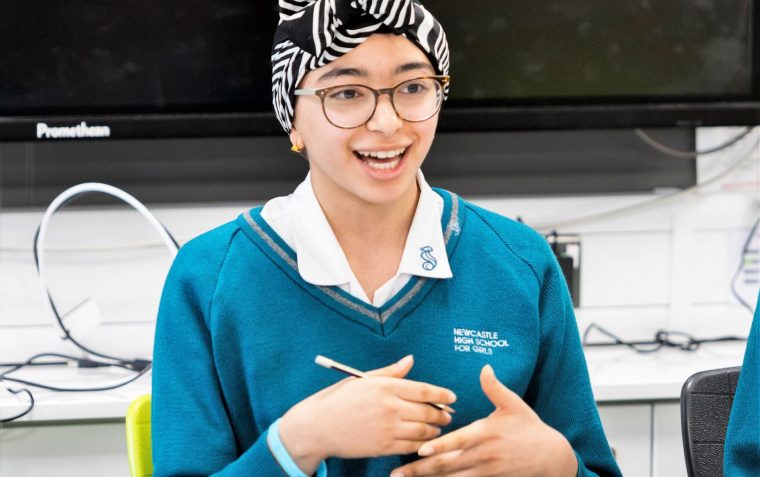 A female secondary school student with head dress and glasses on sitting while speaking with a pencil in her hand.
