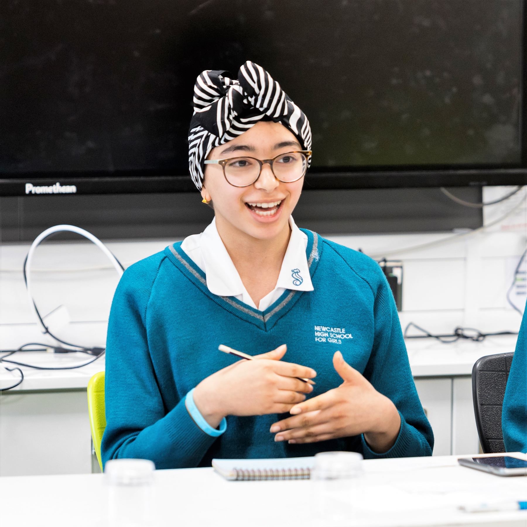 A female secondary school student with head dress and glasses on sitting while speaking with a pencil in her hand.