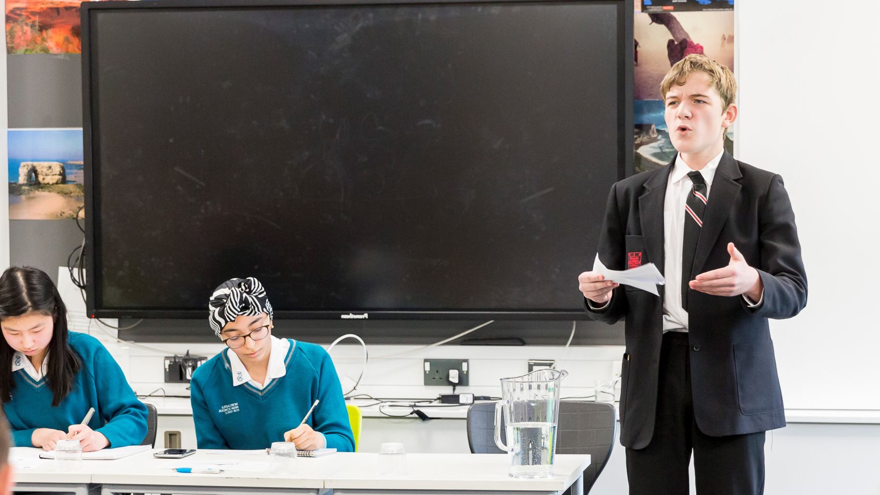 A young pupil in their uniform reading his speech while two other pupils in the background are writing their notes
