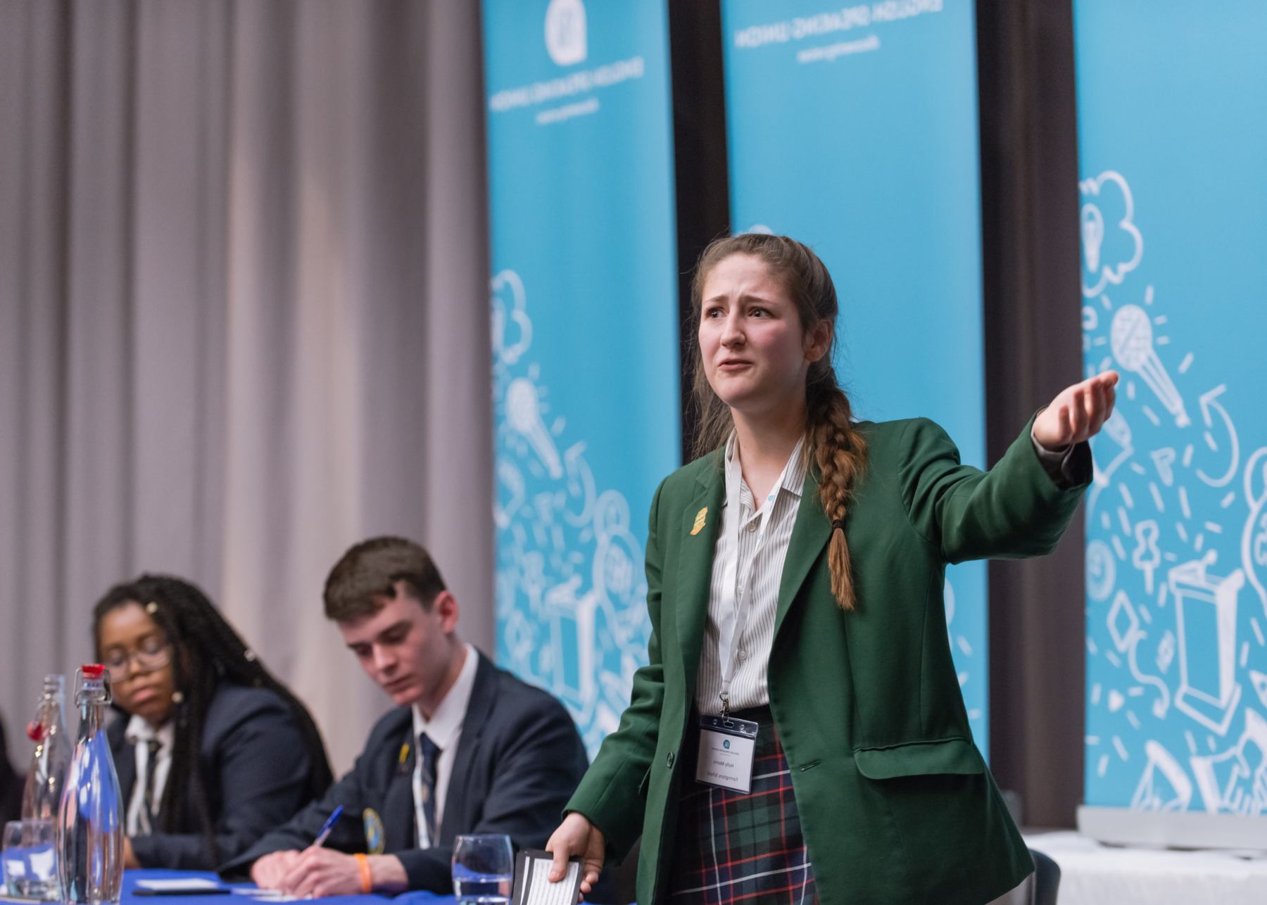 Young pupil in school uniform standing up and speaking on stage, two other pupils are on stage with her