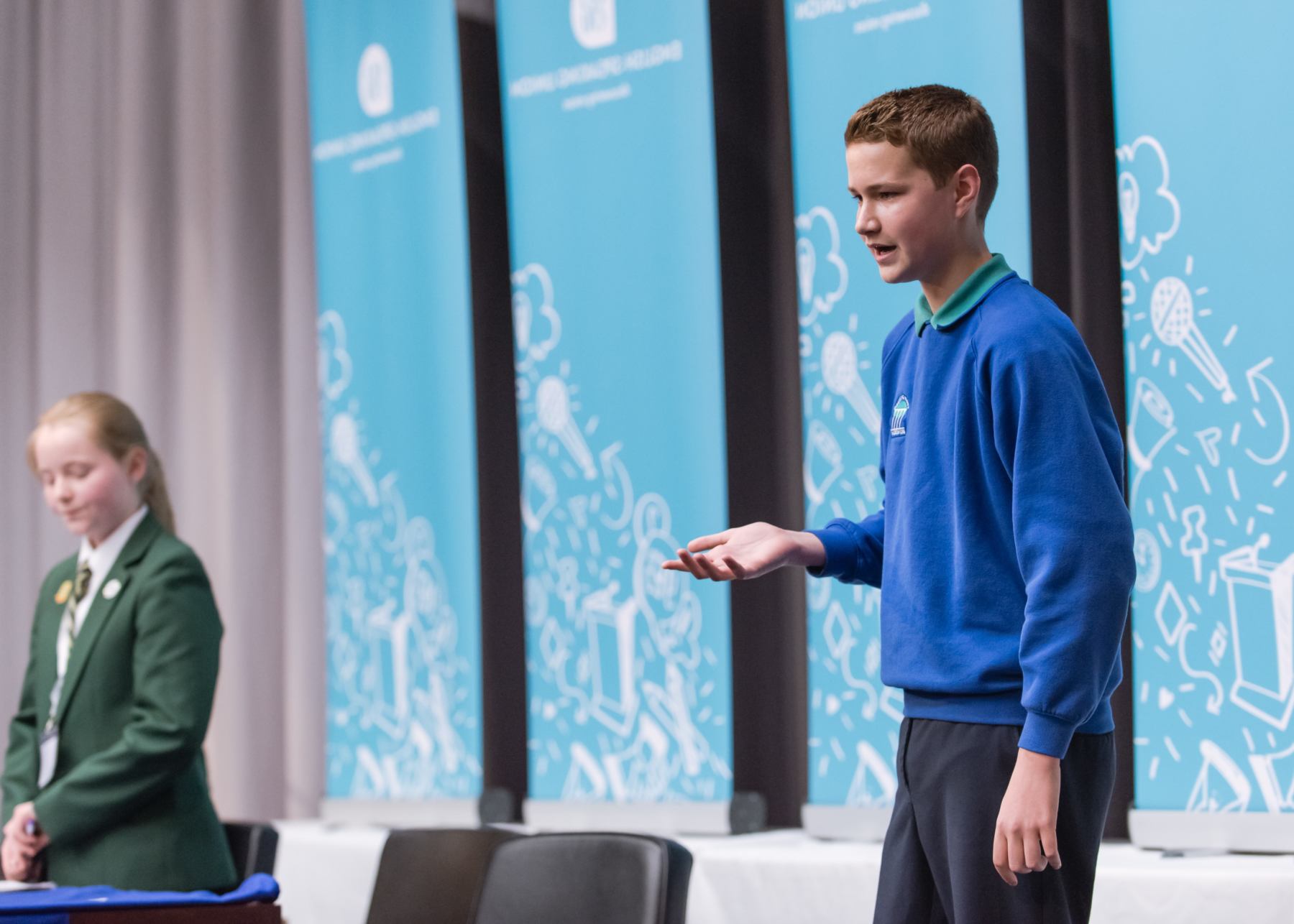 Young pupil in school uniform standing up and speaking on stage, while another pupil is standing and waiting to ask a question