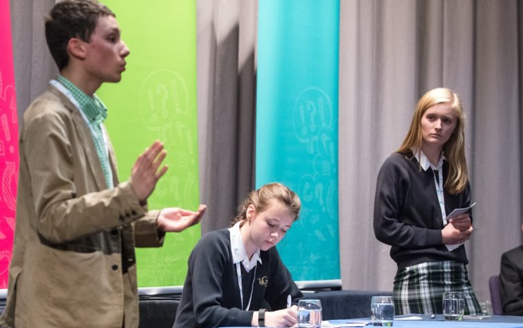 A young pupil giving a speech while another pupil is standing to ask them a question and another pupil is writing her notes